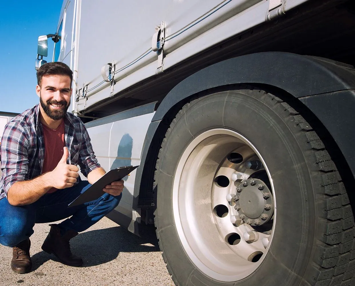 A man is kneeling down next to a truck holding a clipboard and giving a thumbs up.